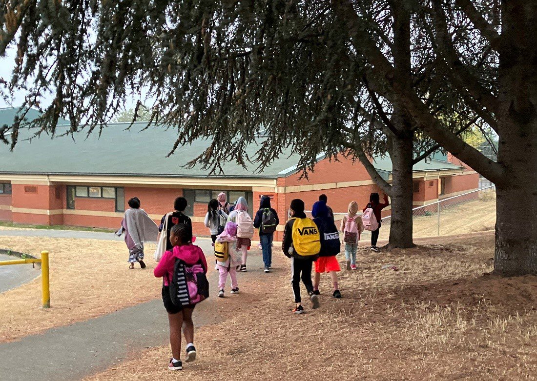 Several young students walk to school. Most students are wearing backpacks, and walking along a path and dried grass, under large trees. The school building is visible in the background.