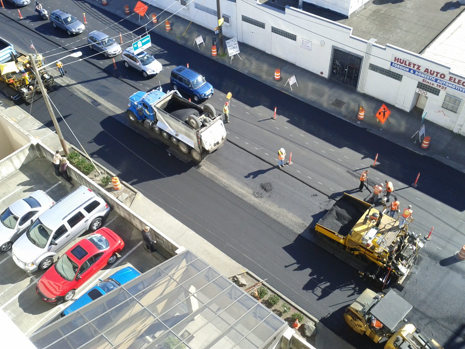 Several large construction vehicles paving the street. Other cars and buildings are in the background.