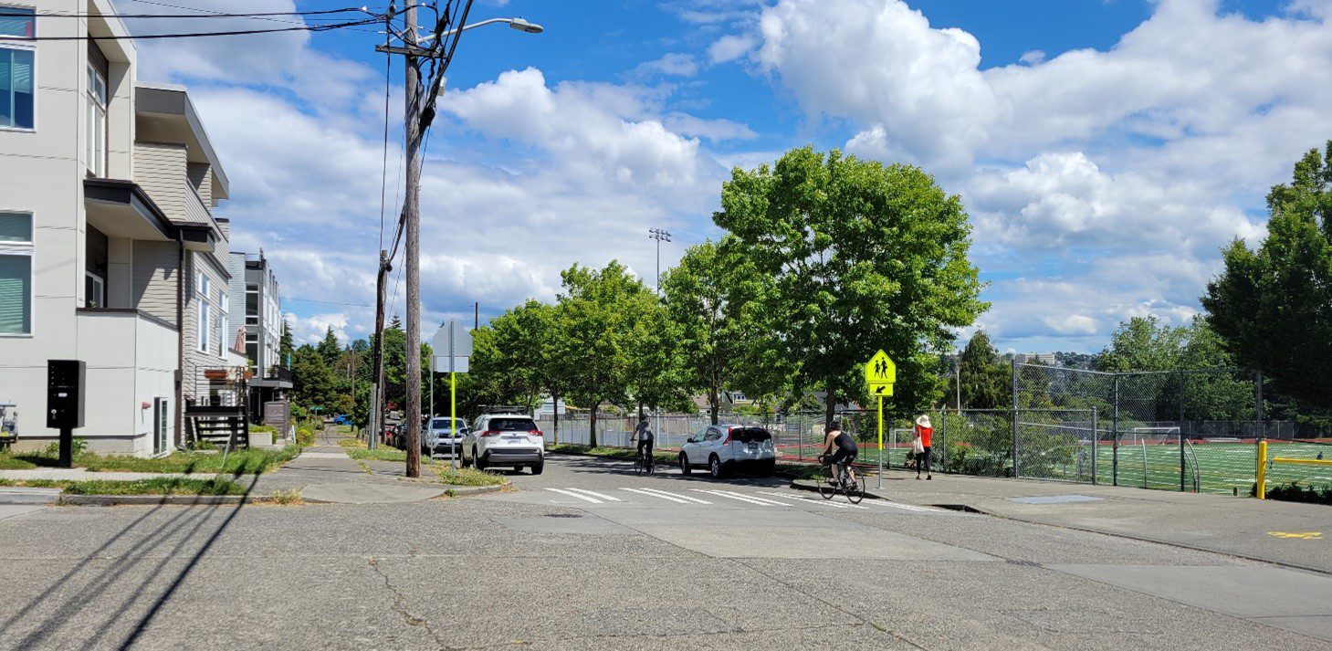 A marked crosswalk on the street, with large mature trees, blue skies, and clouds in the background. A bicyclist travels in the street.