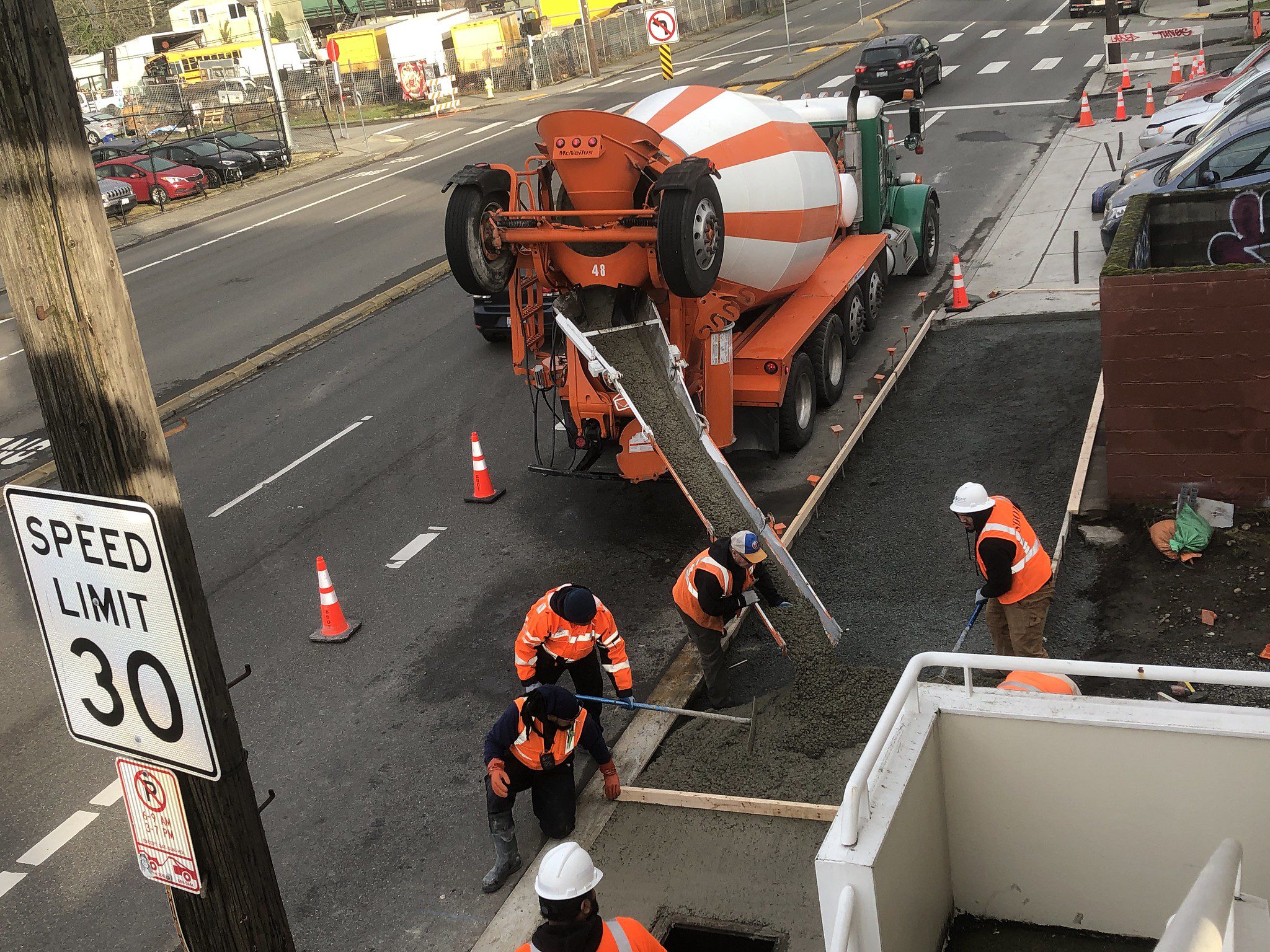 Several people wearing orange construction vests work to pour new concrete along a street. Parked cars are in the background, and a large orange and white cement truck is in the middle of the photo.