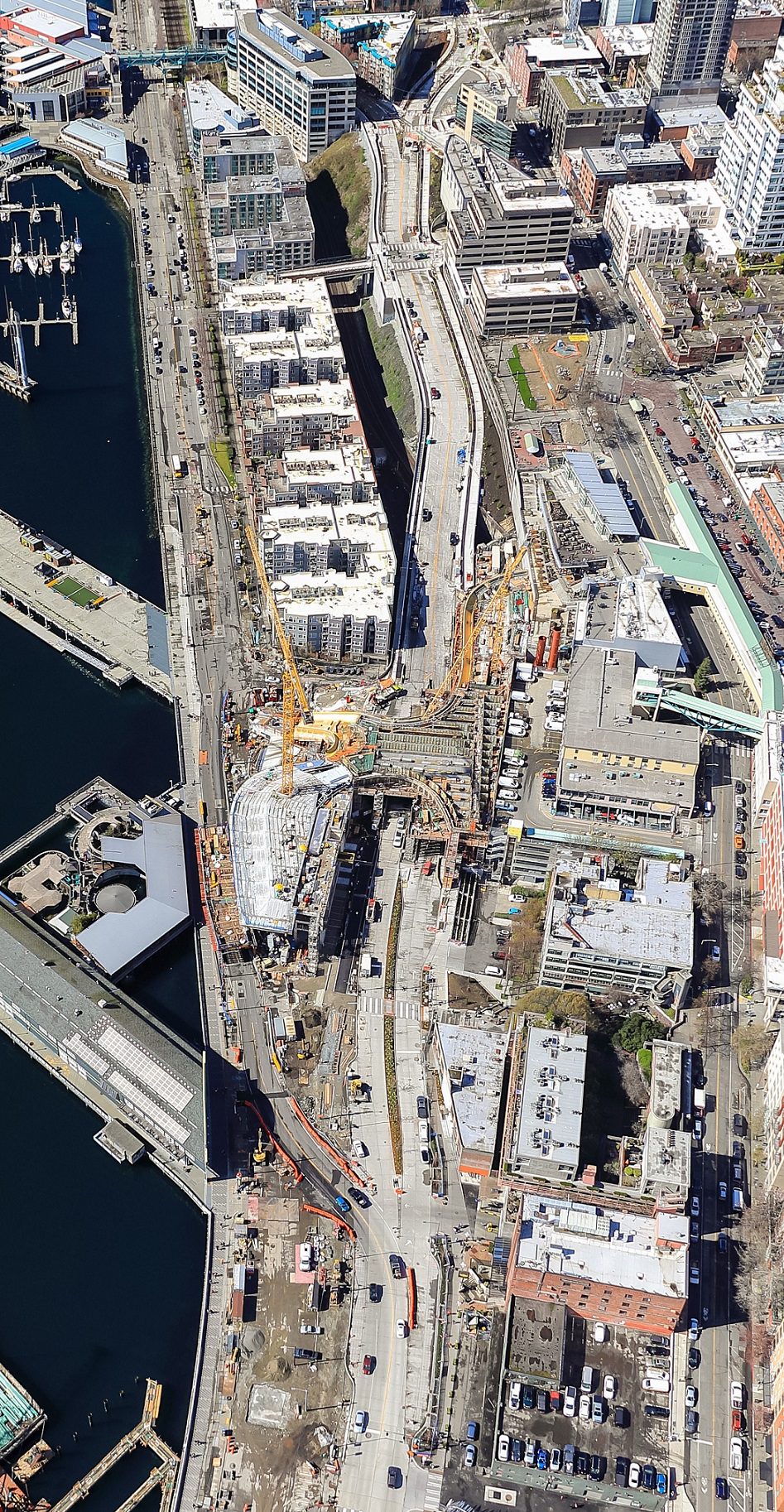 An aerial photo of a waterfront, streets, large buildings, construction cranes, and other cityscape elements.
