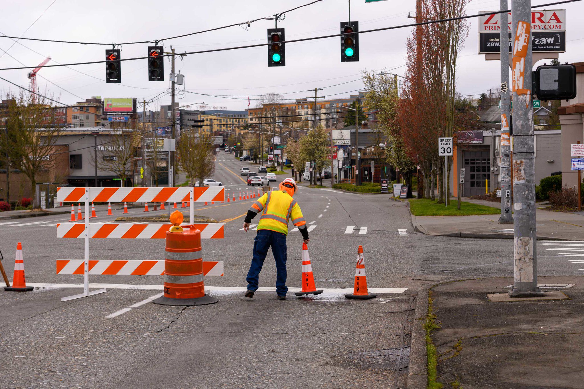 A worker wearing a bright yellow safety vest and hard hat places an orange construction cone in the street at an intersection. An orange and white barricade and orange barrel are also visible in the image. Large buildings are in the background.