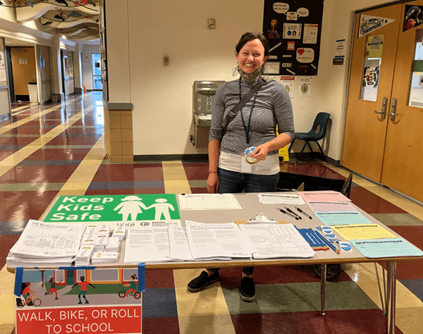 A woman smiles at the camera. She is standing behind a table of informational handouts, inside a school building. Colorful floors, posters, and doors are behind her.