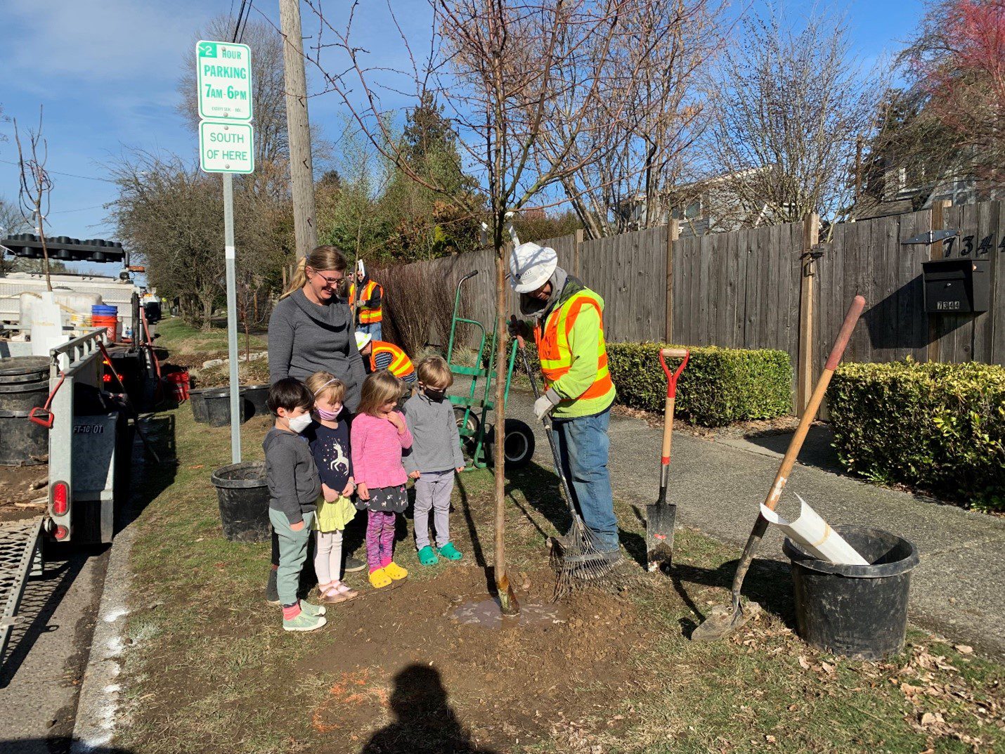Workers wearing orange and yellow safety vests and hardhats work to plant trees in the city. A woman and four kids watch the work taking place. A work truck, tools, hedges, a fence, and large buildings are in the background.