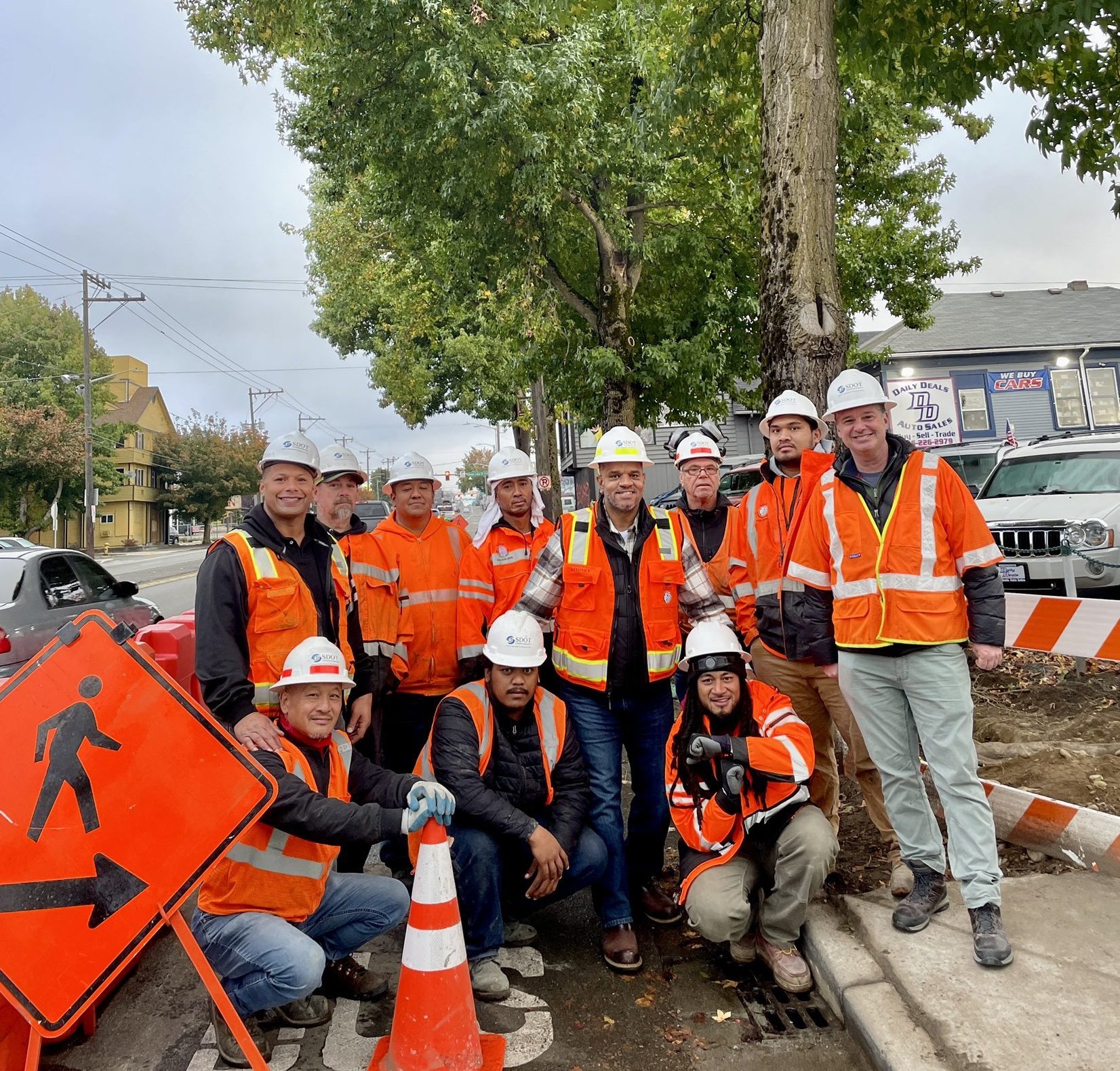 Elevent people wearing safety vests and hardhats smile at the camera near a work zone, with a large tree in the background. Cars and buildings are also in the background.