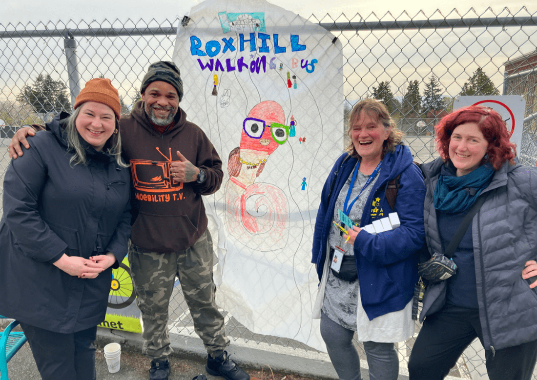 A photo of 4 people smiling while standing on either side of a student-made banner for the Roxhill walking school bus.