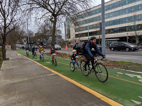 An adult on a bike leads a train of 5 children on bikes down the protected bike lane on 5th Ave.