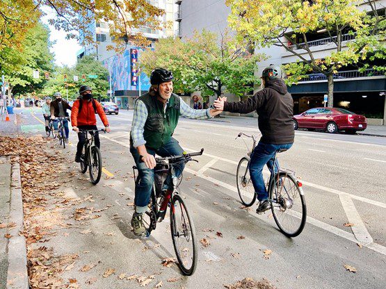 Three riders in the right lane of a downtown protected bike lane exchange high fives with a rider headed in the opposite direction.