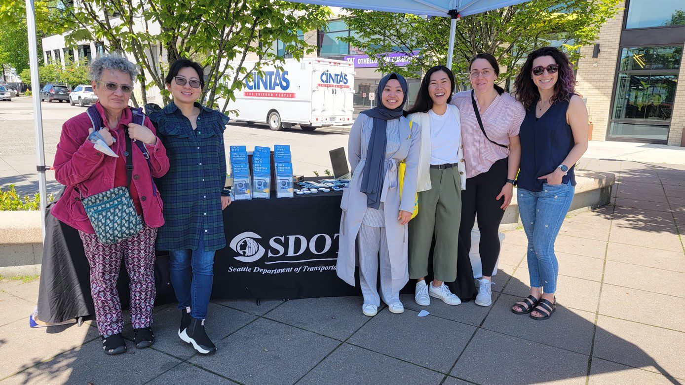 Six members of our TAP and Community Liaison teams pose in a line in front of an SDOT table covered in informational flyers outside the UW Othello Commons.