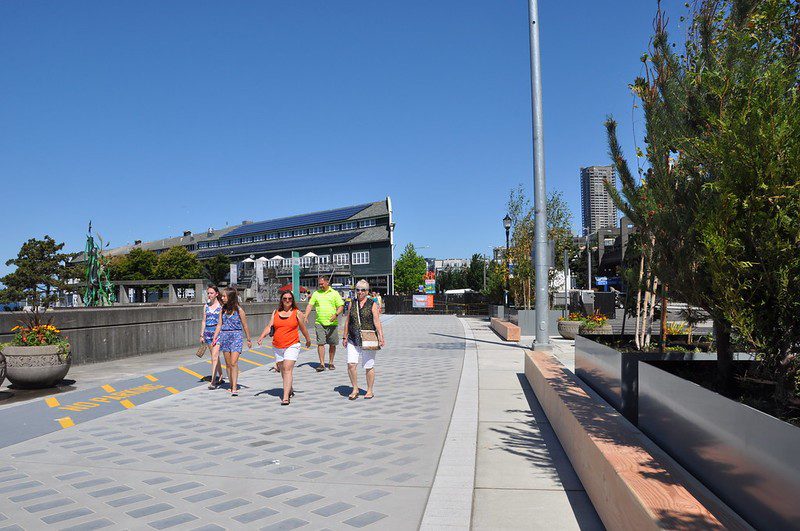 A group of 5 pedestrians walks along the waterfront on a warm, sunny day. They face the camera, and the Seattle Aquarium building is visible behind them.