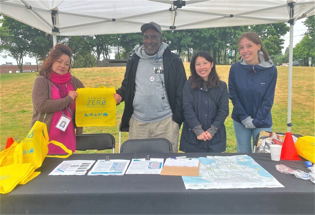 4 members of our staff stand behind the SDOT event table and smile at the camera. Two of them hold up a Vision Zero tote back between them.
