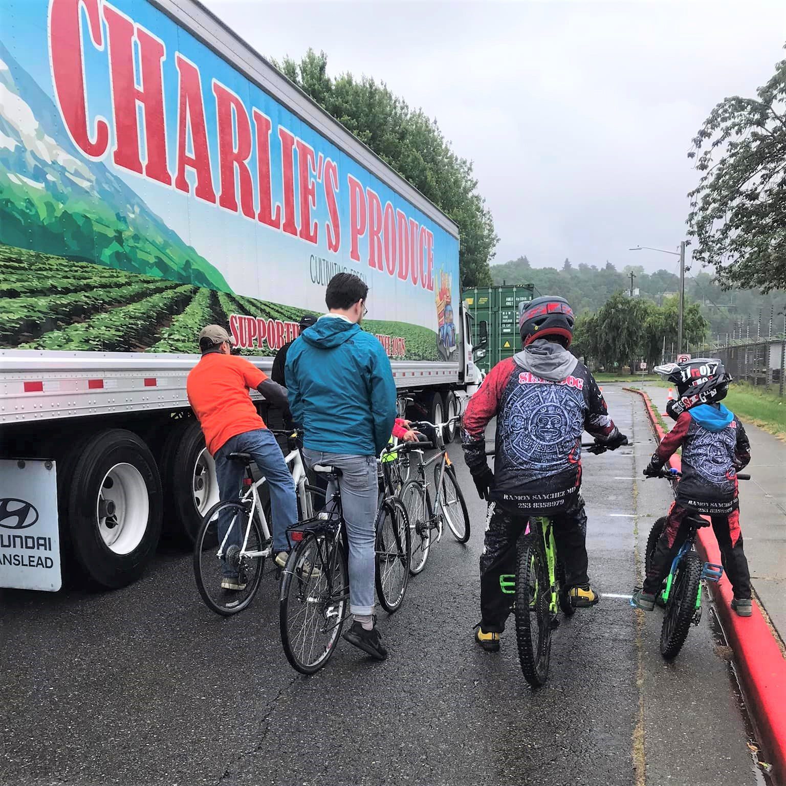 4 adults and one youth sit on bikes on the street between a Charlie's Produce semi truck and the curb. They are discussing the trucks blind spots. 