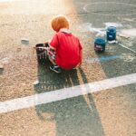 A small boy in a red shirt crouches on asphalt next to a bucket of sidewalk chalk sticks. He colors on the roadway with his right hand with his back to the camera and the sun shining on his head.