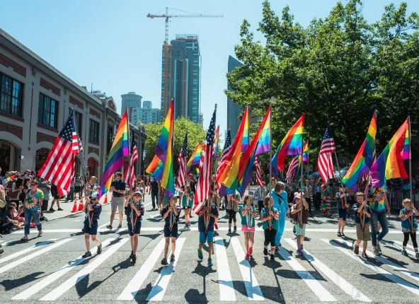 A group of young people walk side by side in a line down the middle of the street downtown, each carrying a large rainbow flag or American flag.