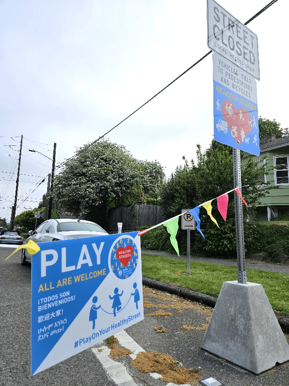 A Healthy Street closure sign in a triangular concrete planter sits in the right shoulder of a road with a colorful string of plastic flags tied to the pole and extending out past the left side of the image. Hanging in the middle of the flag banner is a new Play on Your Healthy Street sign.