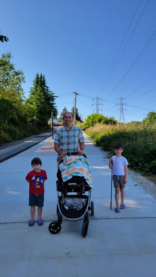 A picture of a father pushing a stroller on the new sidewalk. He is with two of his sons who stand on either side of the stroller.