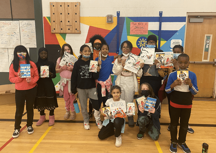 A group of 12 elementary students stand in their school gym and pose for the camera with their favorite books with walking and biking themes.