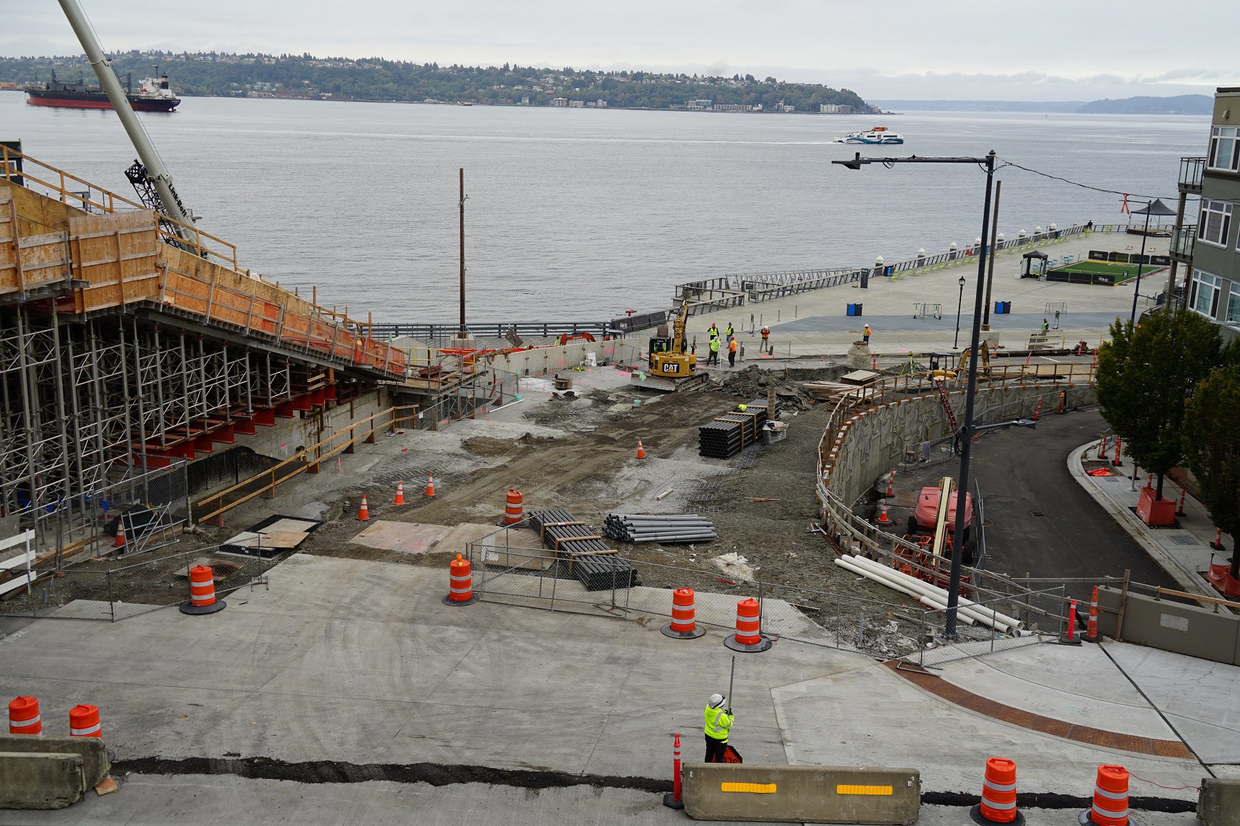 View looking down a construction site toward a large body of water on a cloudy day. Several large orange barrels and construction workers are visible.