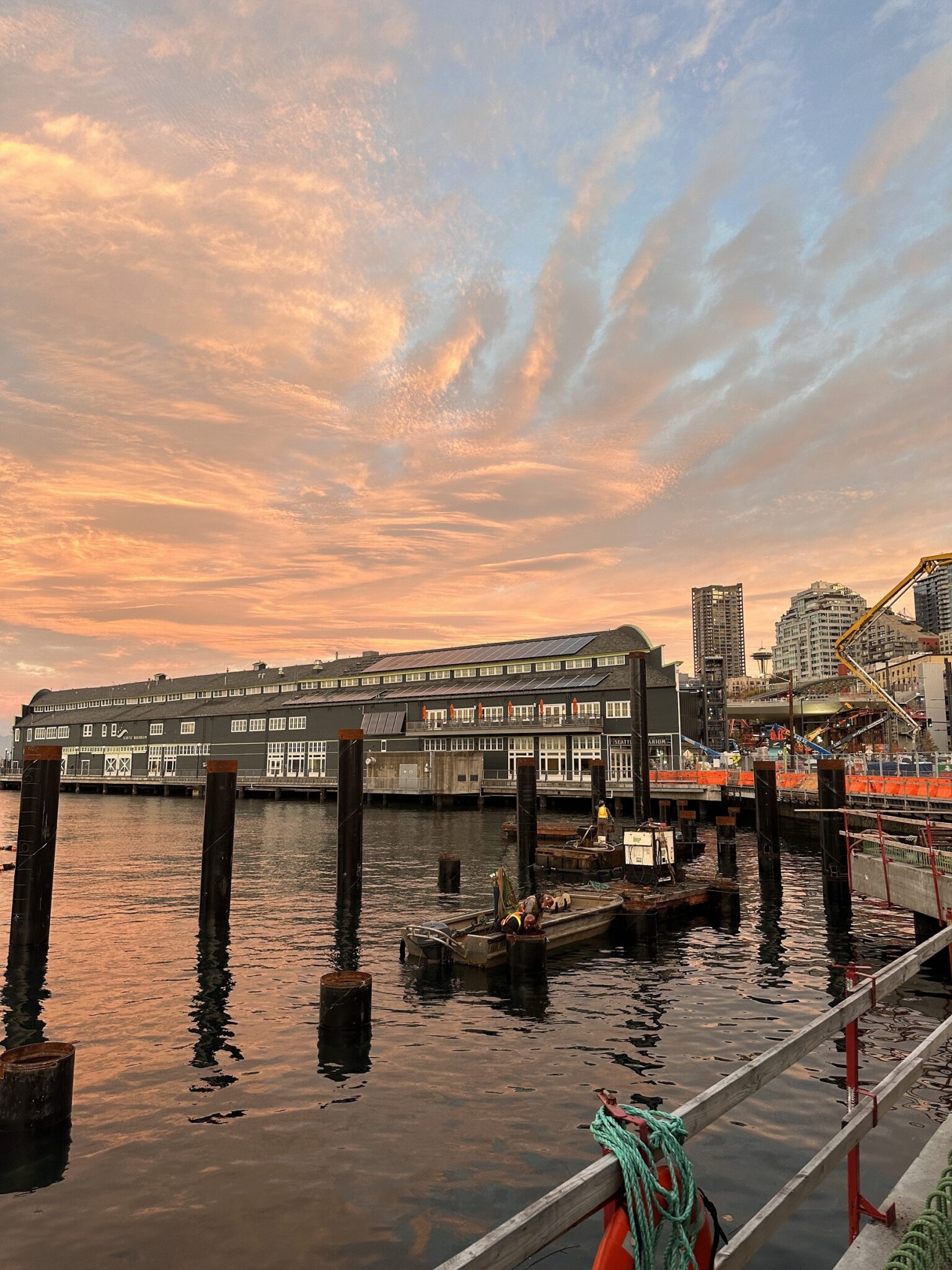 Photo of construction along water near sunset. Large buildings are in the background and middle of the image. A small boat is in the middle in the water.