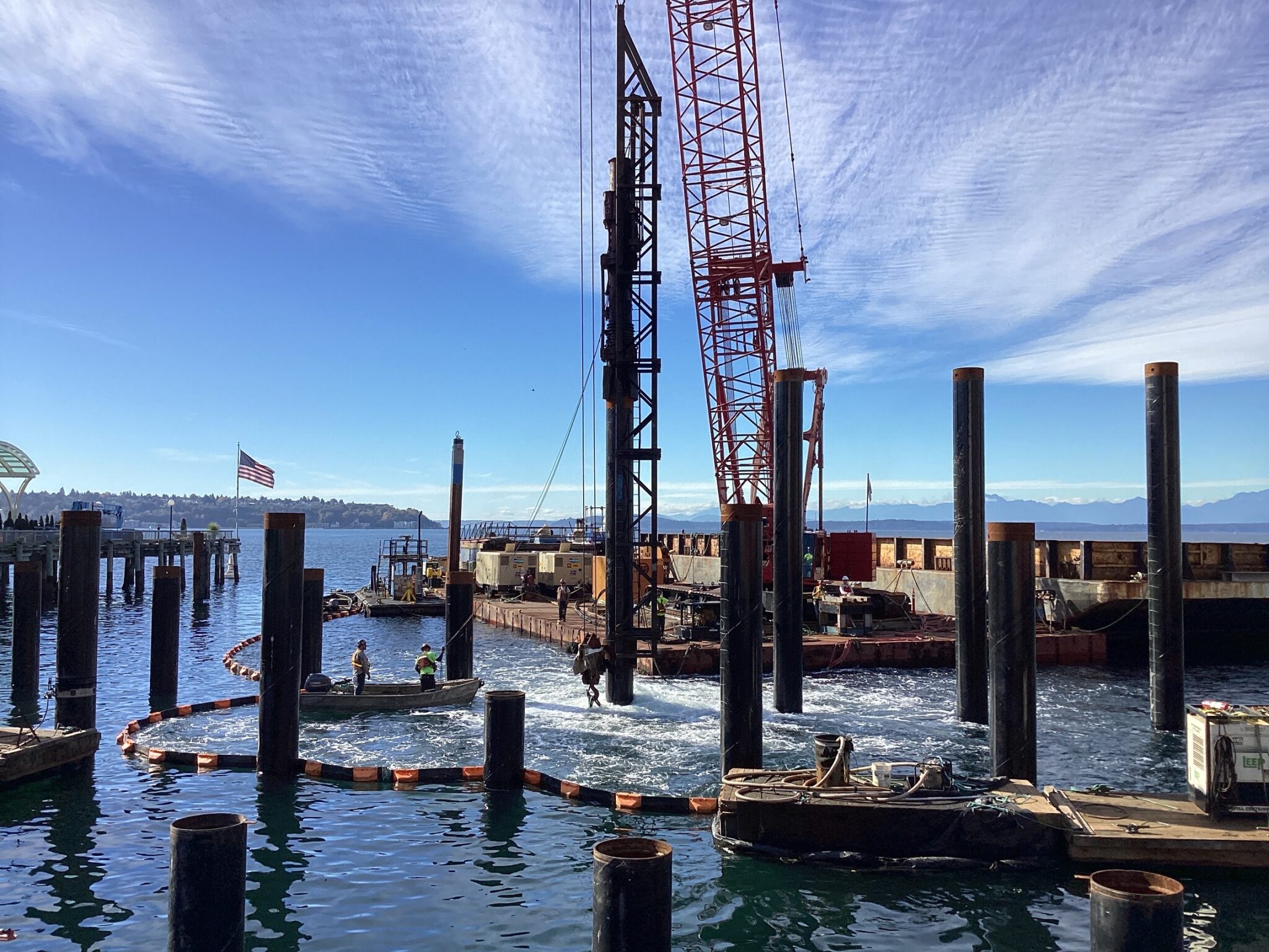 Image of pile installation at a large body of water on a sunny day. Work crews and a large crane are in the background.