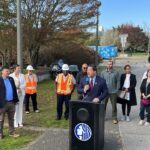 Mayor Bruce Harrell stands at a podium outdoors at Sam Smith Park to address the crowd gathered for the groundbreaking. Sound Transit CEO Julie Timm and SDOT Director Greg Spotts stand behind him.