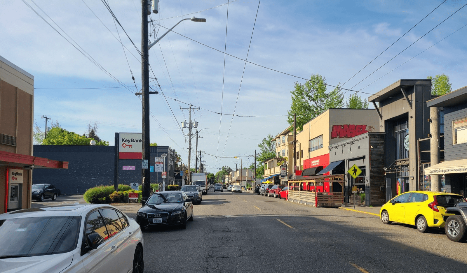 A photo of 15th Ave E in the middle of a week day, with parked cars visible on both sides of the street, with a street cafe taking up a portion of the curb space to the right, immediately outside of the restaurant Smith.