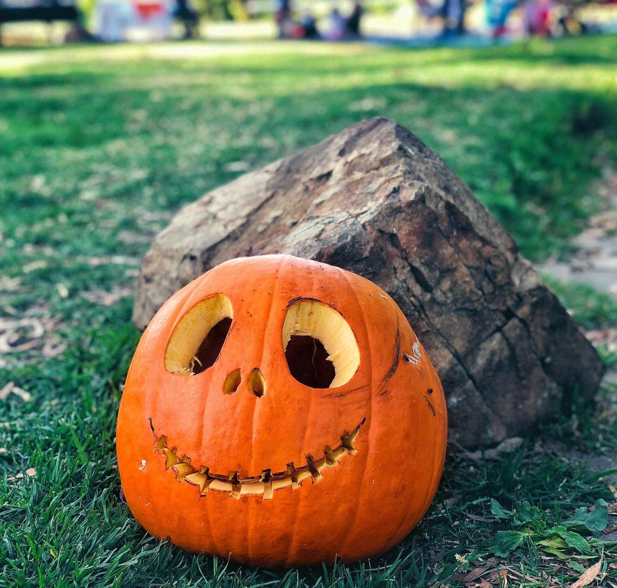 A smiling jack-o-lantern sits on the grass near an outdoor Halloween party