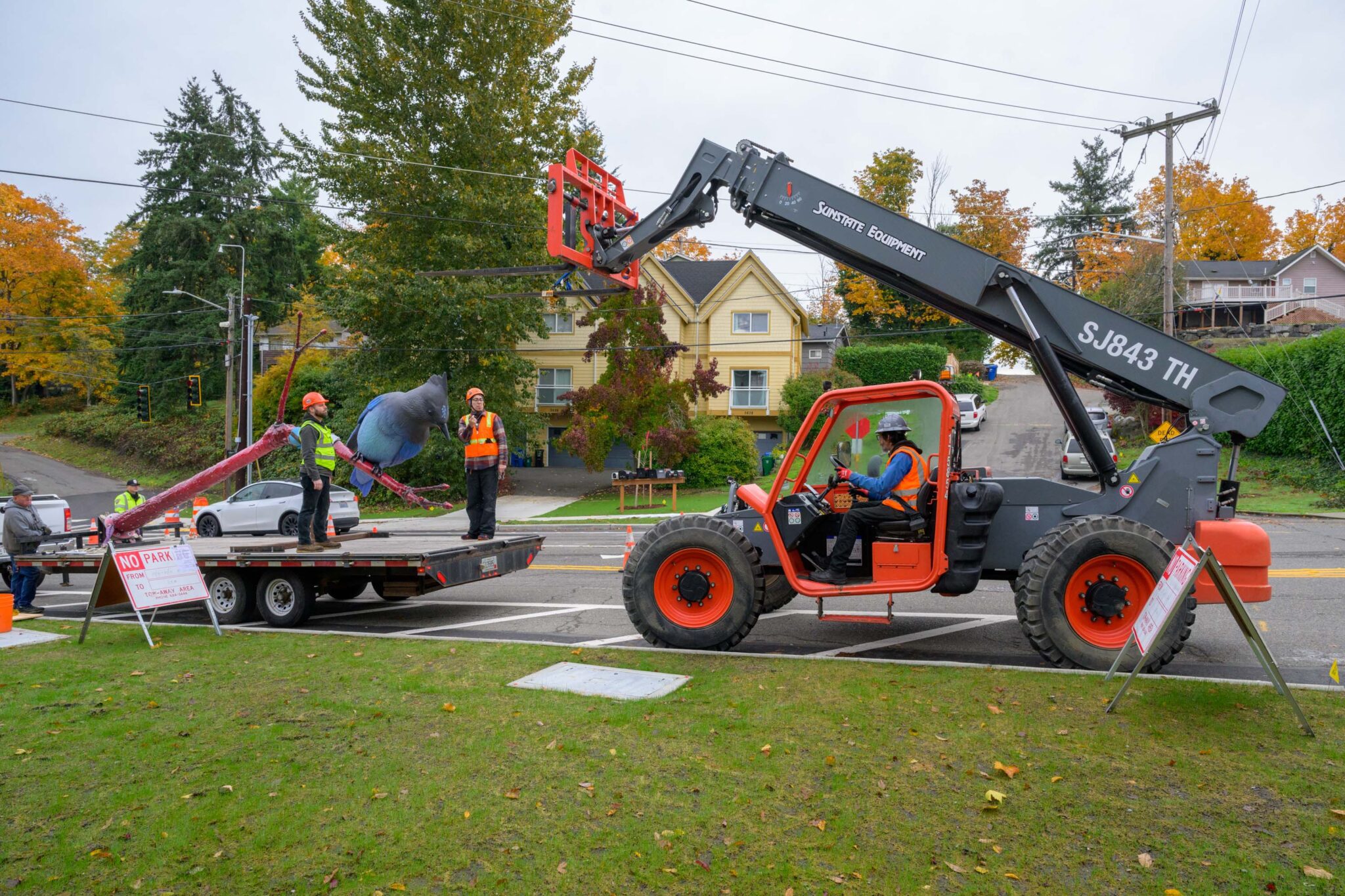 Workers use large heavy equipment to unload the art near a large grassy area with large trees and houses in the background.