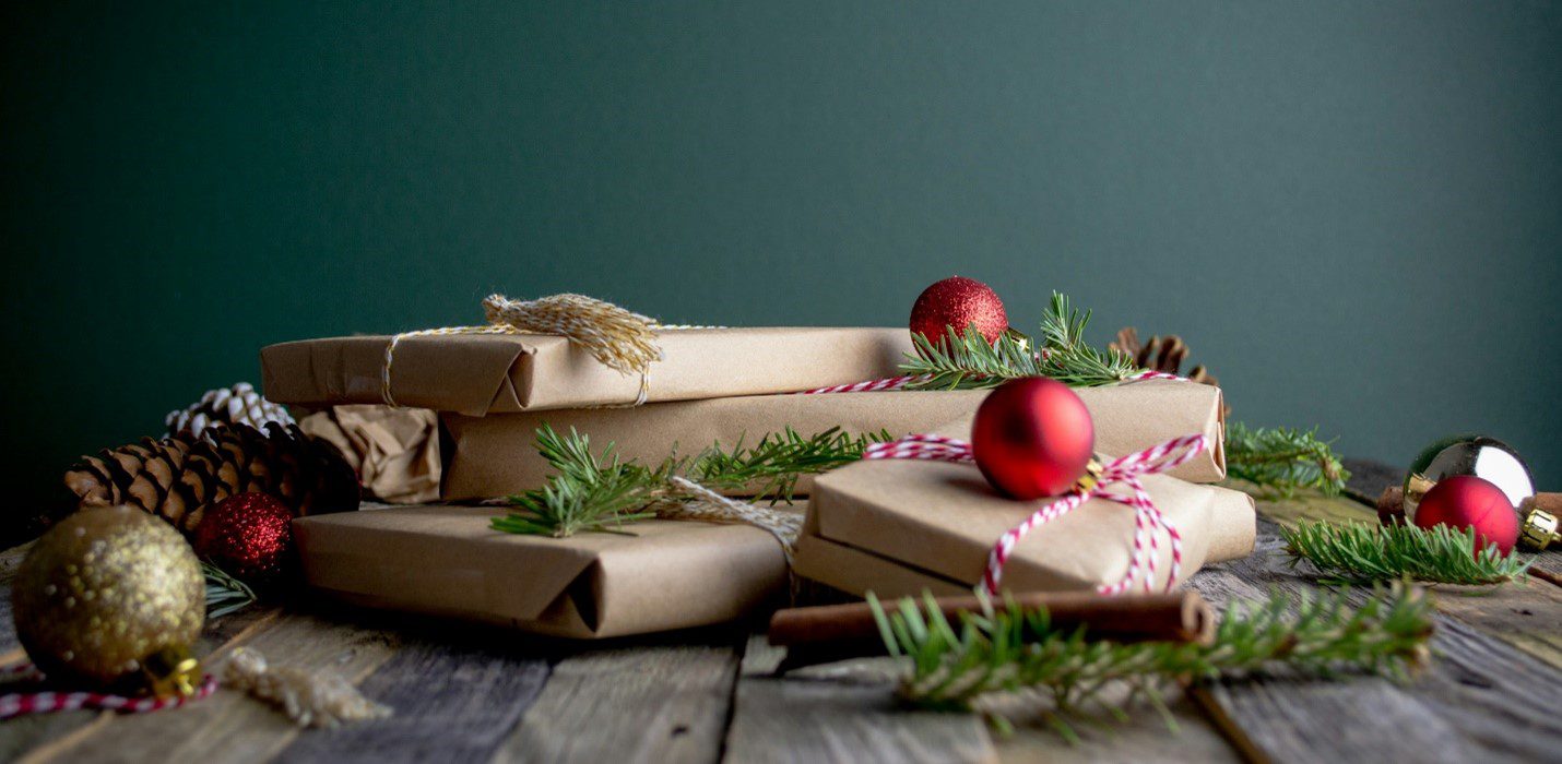 Several packages wrapped in brown paper on a table with holiday decorations on top, including red orbs and pine needles.