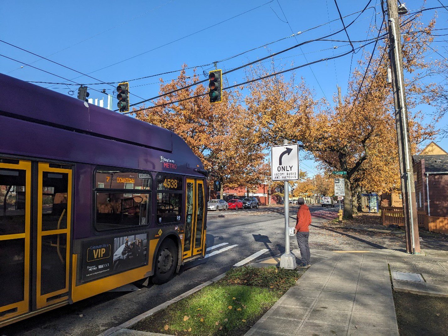 A large bus travels on a street with a person standing on a sidewalk nearby. Blue skies and large trees are in the background.