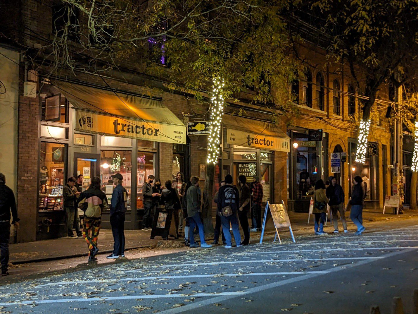 People standing outside a music venue on a street in the nighttime. Large buildings, trees, and no parking signs are in the image, as well as lights on the trees.