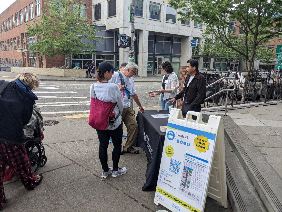 People stand at an outreach table and community members speak with project staff. Large buildings, parked cars, and people are visible throughout the photo.