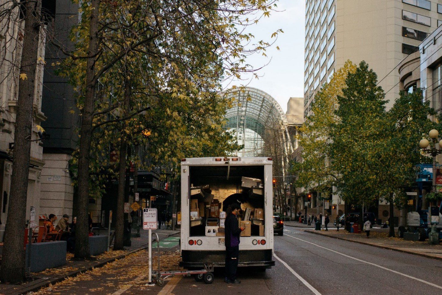 A worker delivers packages from a large white delivery truck. Trees and large buildings are in the background, as well as a street and sidewalk.