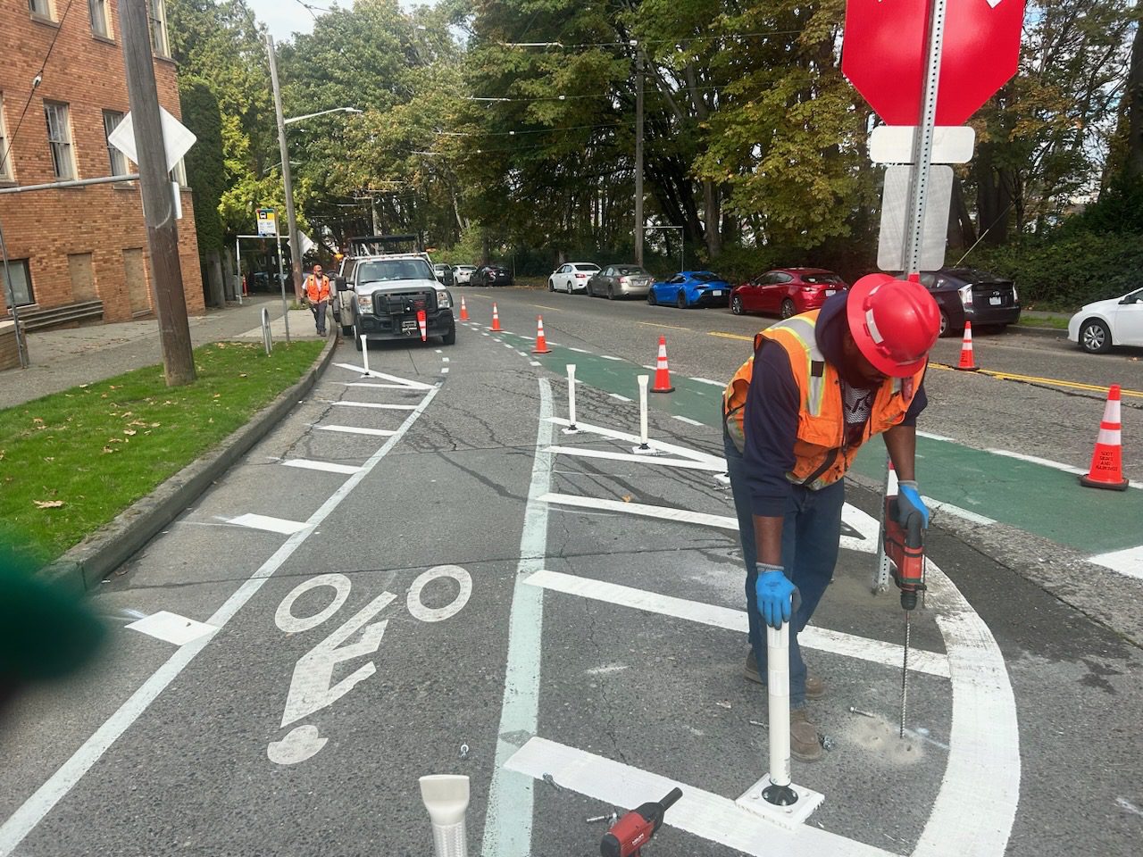 A worker wearing an orange vest and red hard hat installed flex posts as part of a new all-way stop, next to a marked bike route. Parked cars are in the background.