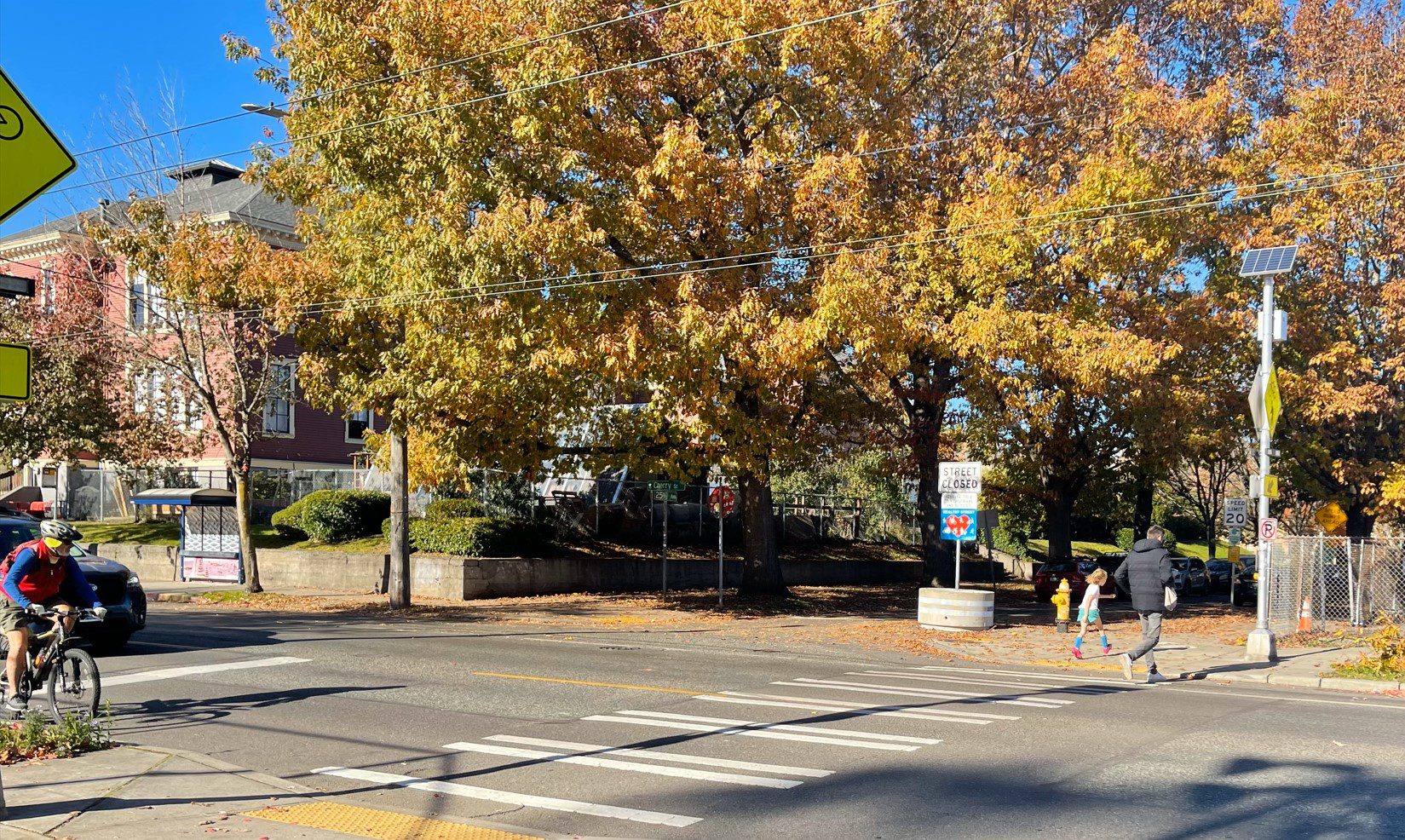 People cross the street while a person biking and person driving wait to pass, on a crisp sunny fall day.