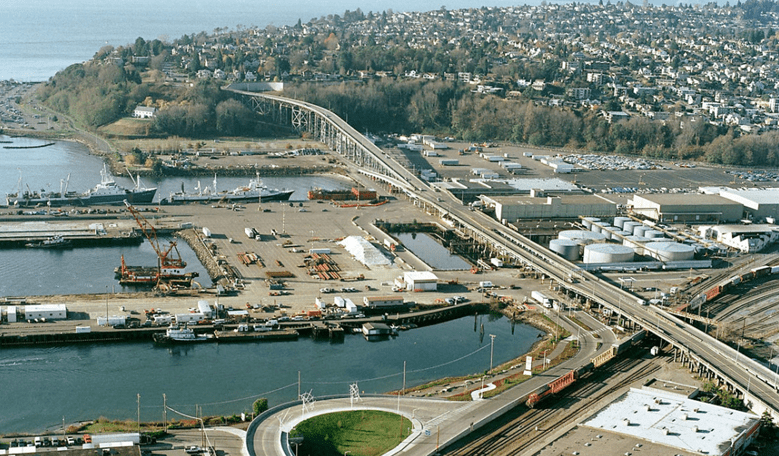 Aerial view of a large bridge with houses in the background on a hillside, water, and industrial area in the foreground.