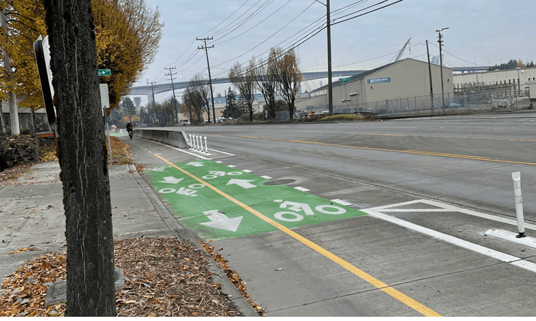 View of a protected bike lane with green paint marking where it crosses a driveway. Vehicle travel lanes are to the right, with large trees and the West Seattle Bridge in the background.