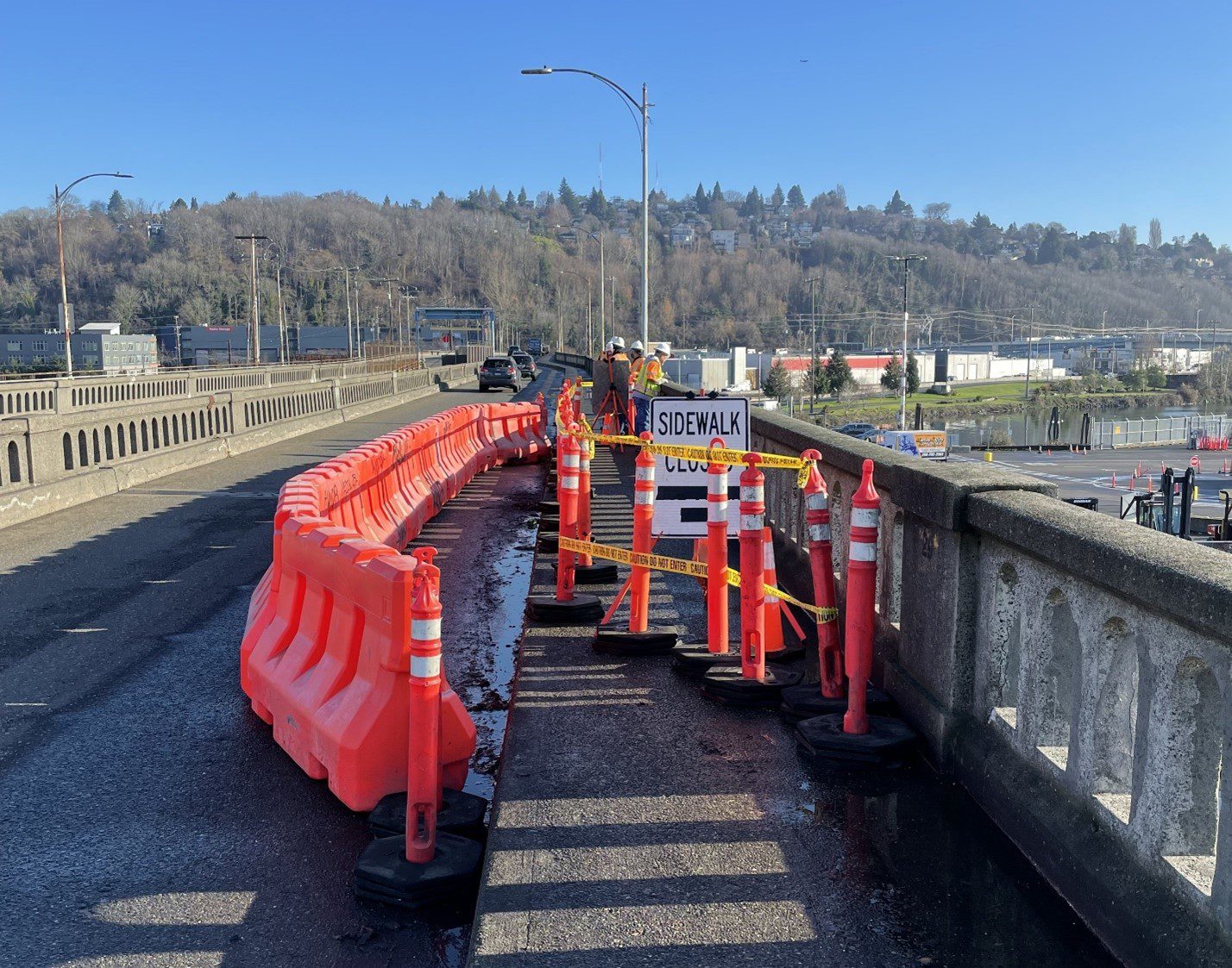Work crews wearing hard hats and high visibility vests work atop a large bridge. Orange cones and barrels show where a sidewalk is closed and a temporary pathway has been created.