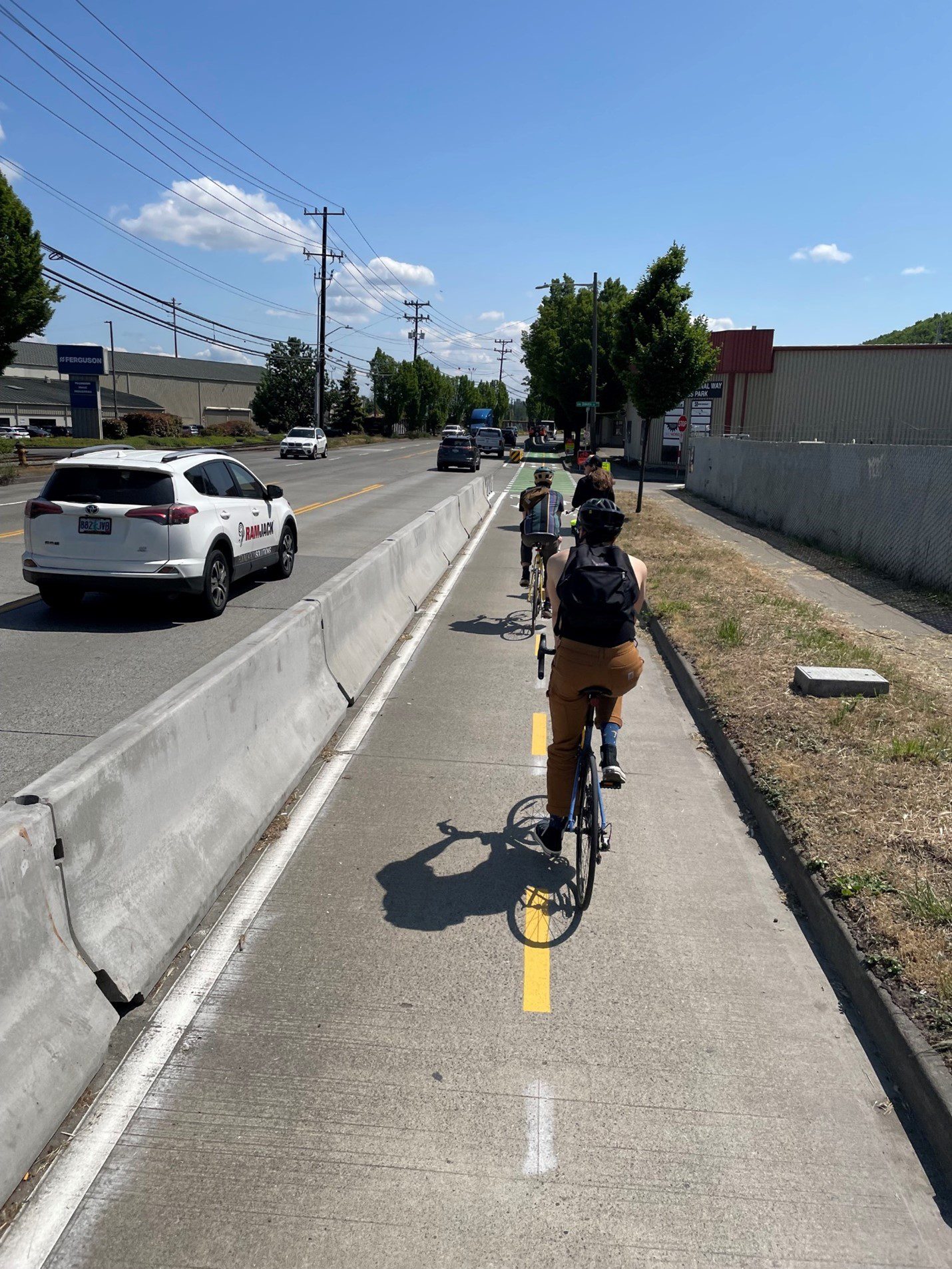 Several people bicycle in the protected bike lane along West Marginal Way SW. Cars travel in the street travel lanes to the left.