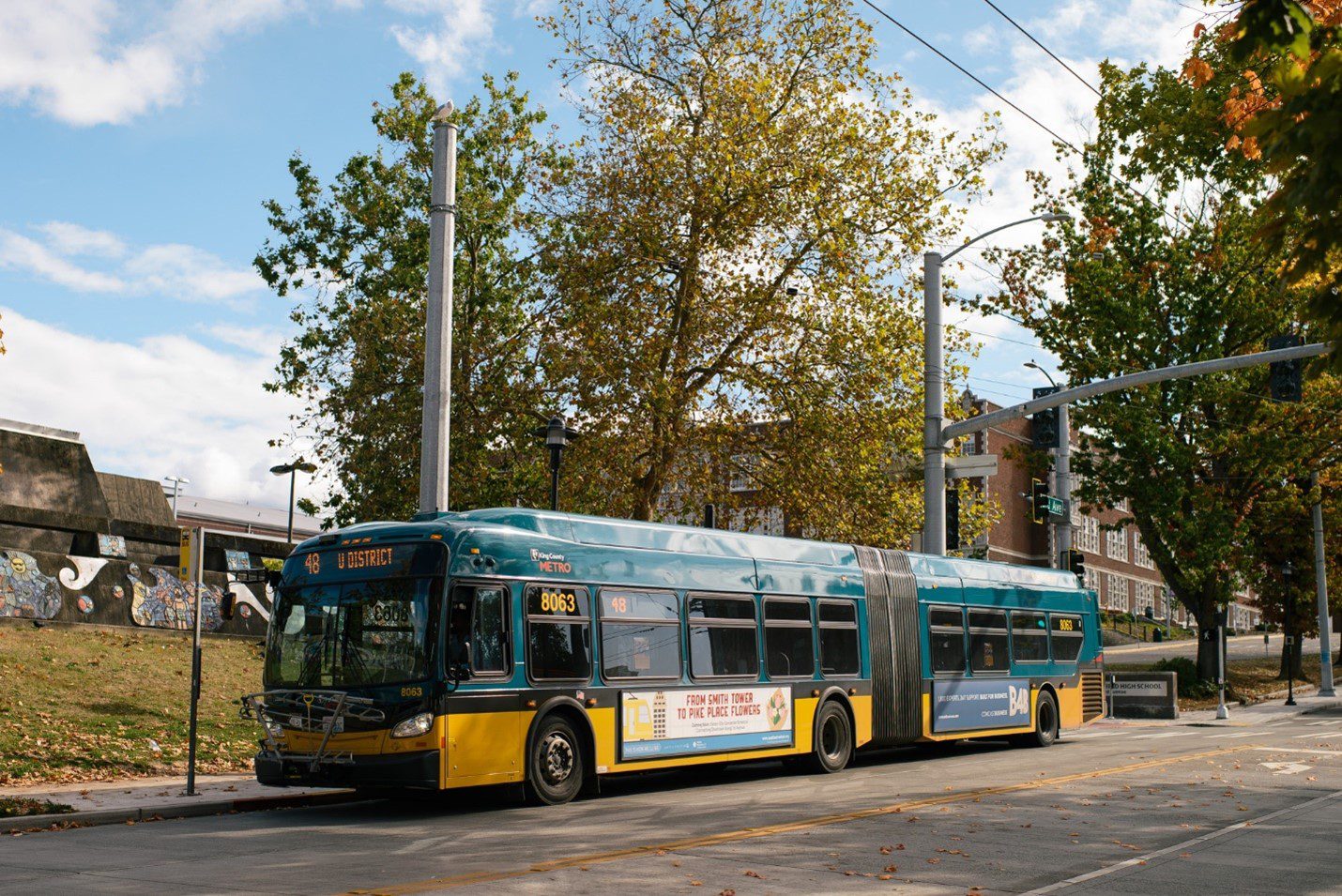 A King County Metro bus number 48 waits at a bus stop on a partly sunny fall day. Large buildings and mature trees are in the background.