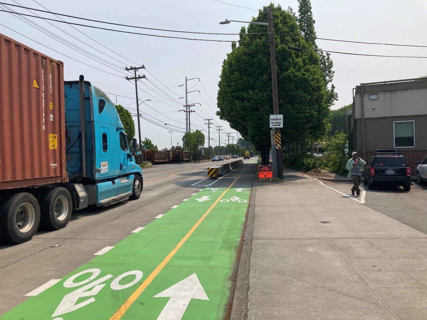 A large semi-truck hauling freight cargo travels to the left of the photo. A green protected bike lane at a driveway crossing is in the middle of the photo. A man walks at the sidewalk to the right of the photo next to parked cars and a building. Large trees are in the background.