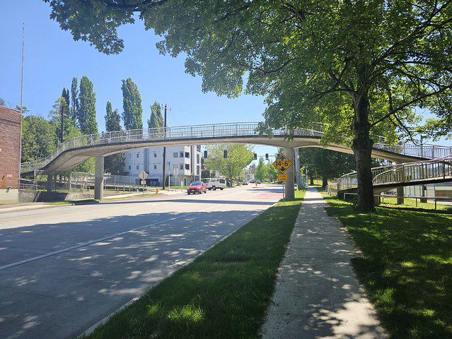Photo of a pedestrian bridge crossing over a street on a sunny day. Cars are in the background, and grass and a large tree are to the right.
