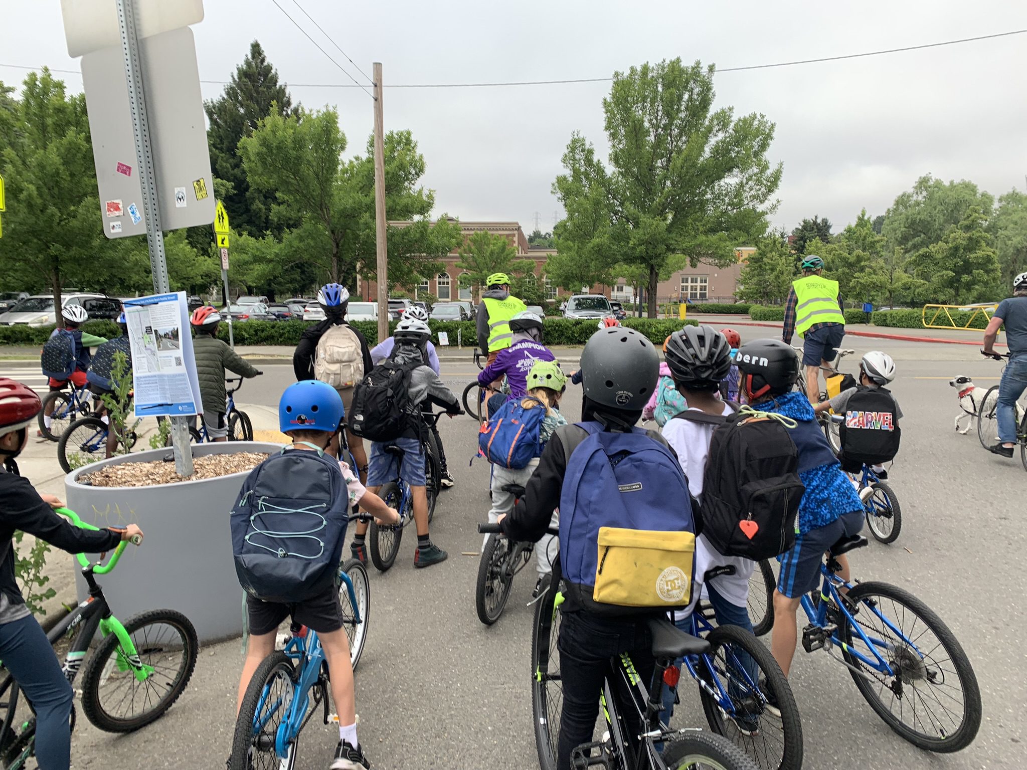 Photo of many students riding their bikes to school in Seattle on a partly sunny day. The school building and trees are in the background.