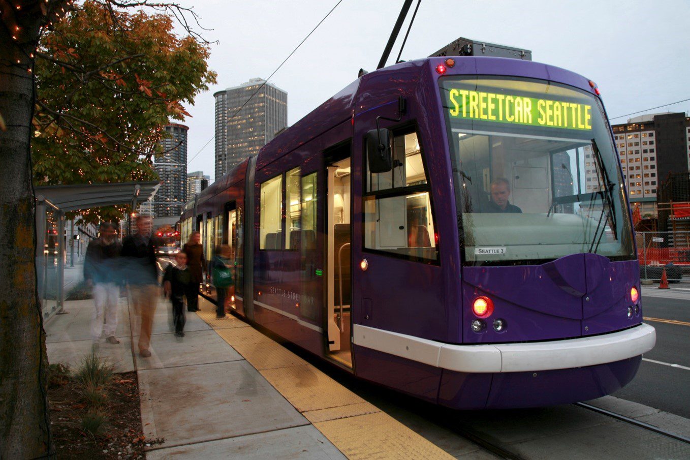 Image of a purple streetcar at a stop with people walking nearby.