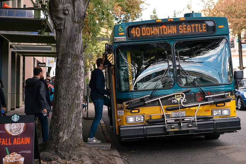 A Route 40 bus en route to downtown Seattle picks up passengers from a sidewalk area. A large tree is to the left.