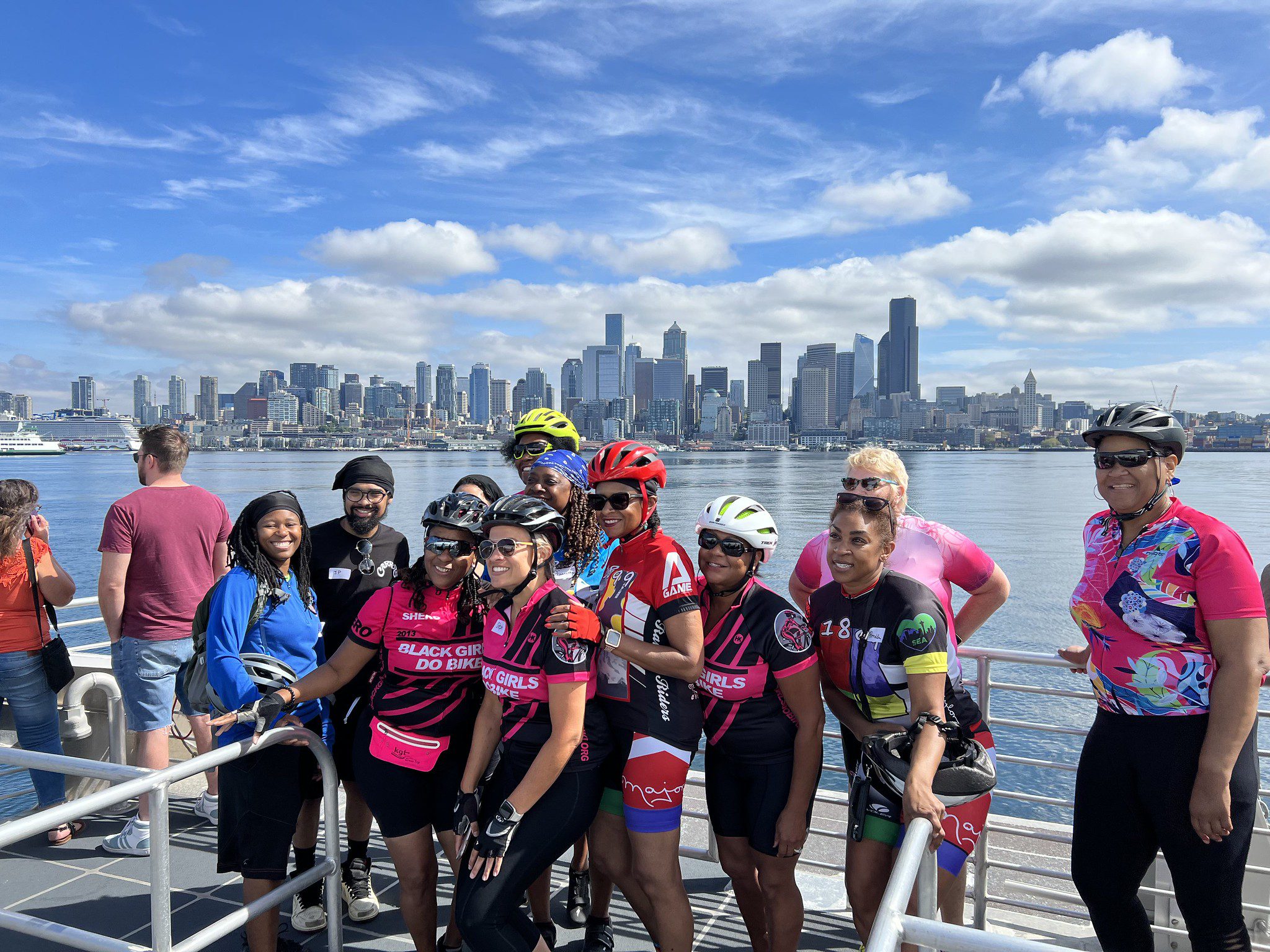Several Black women and community members smile while riding on a boat with a cityscape in the background on a sunny day. Most people are wearing bike gear and several people are wearing bike helmets.