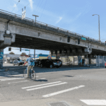 A person bikes across a crosswalk on a sunny day in front of a large bridge.