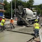 Several construction workers installing concrete pavement on a street. A large cement mixer truck in the center of the image.