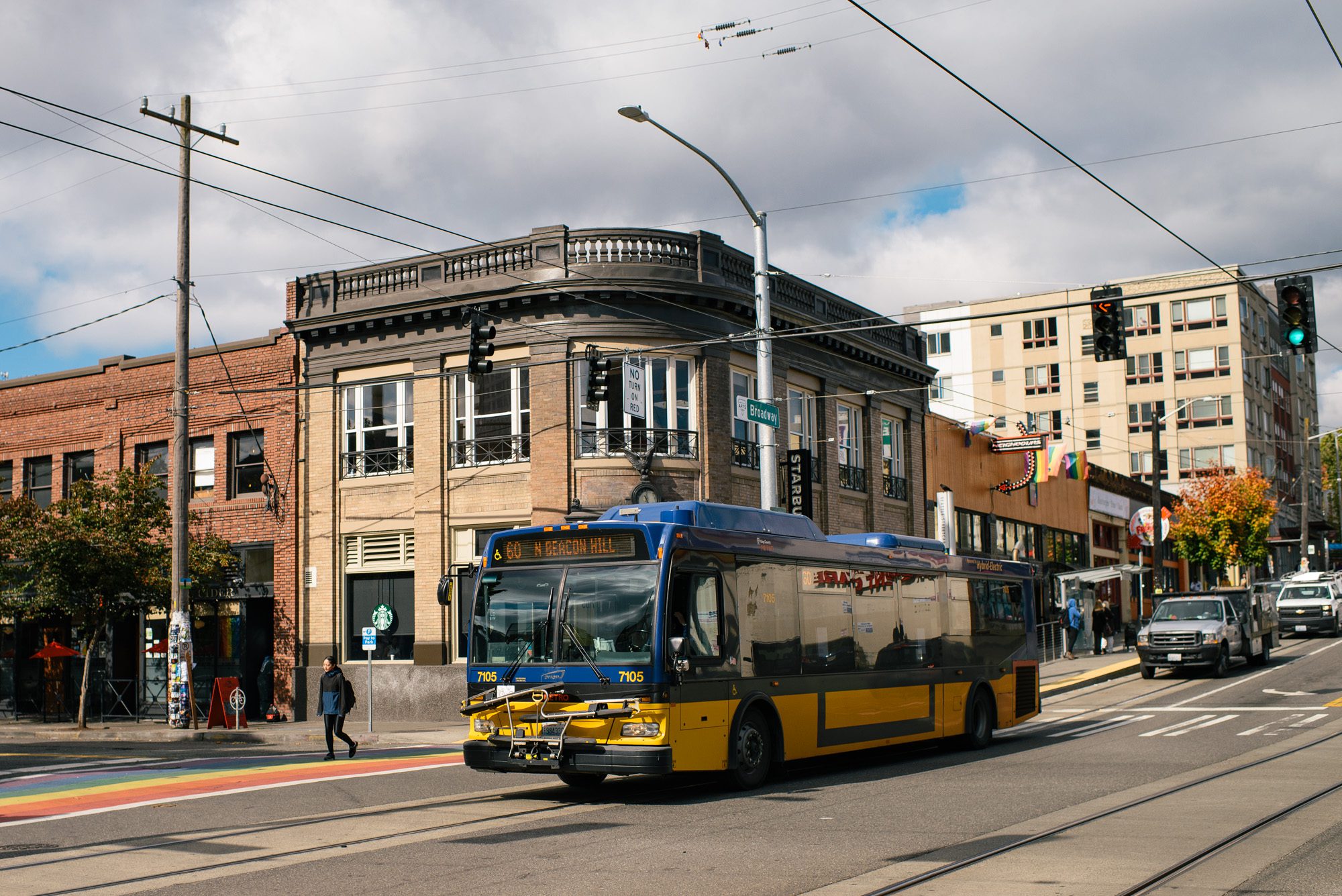 A Route 60 bus to north Beacon Hill travels down the street on a partly cloudy, partly sunny day. A person walking crosses the street to the left, while two trucks travel in the street behind the bus.