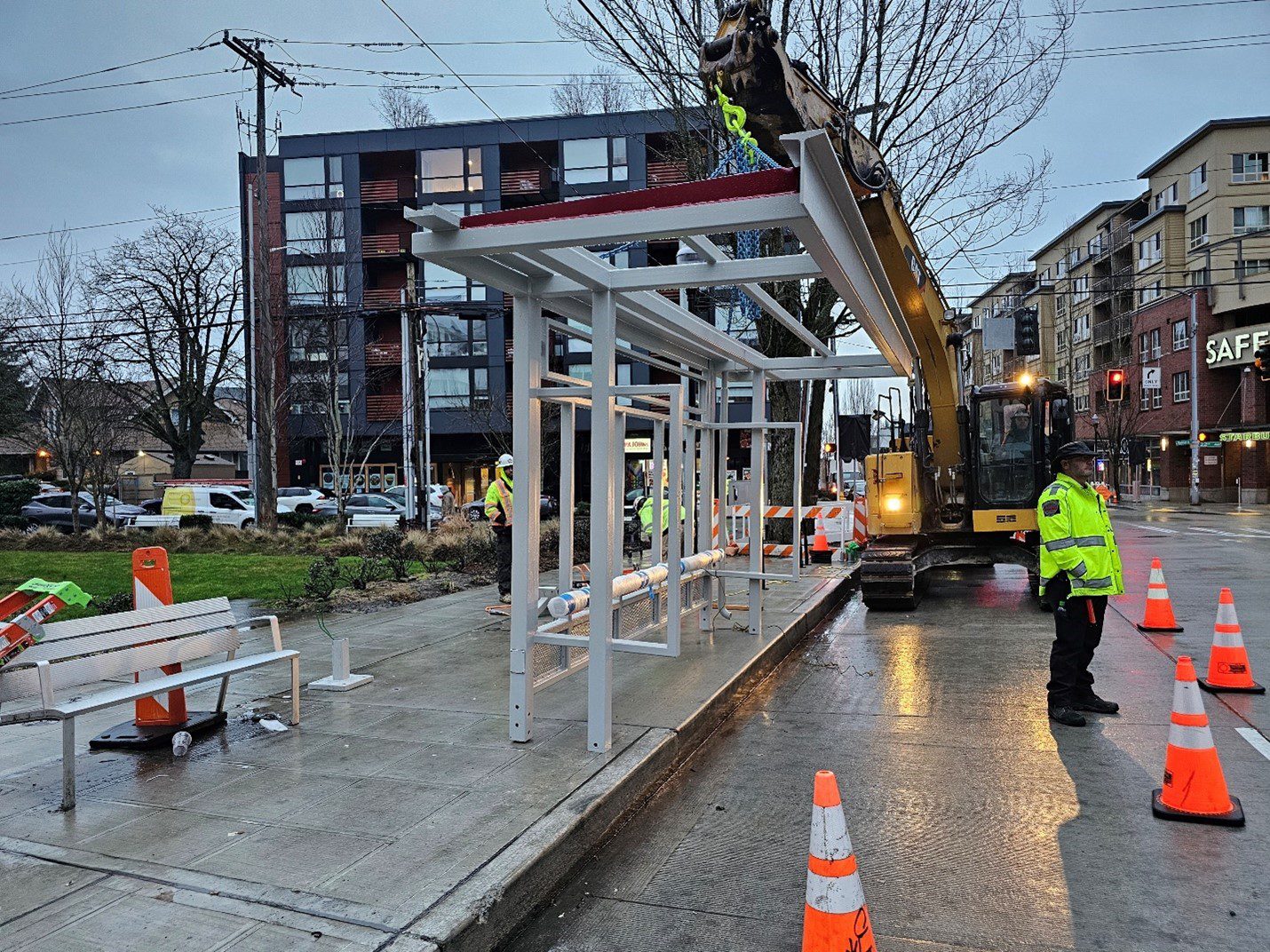 Crews working to install a new bus shelter with heavy equipment. Construction workers and a traffic officer stand next to the installation work. Large buildings are in the background.